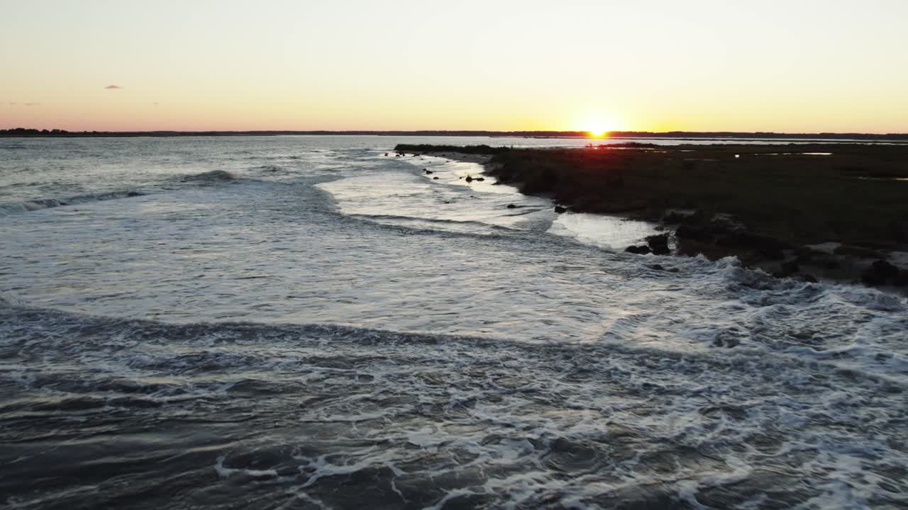 Onde dell'oceano si infrangono lungo le coste sabbiose in pendenza mentre il sole tramonta sotto l'orizzonte con un bagliore sfumato giallo-blu nel cielo
