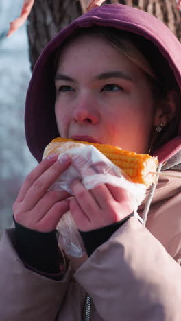 joven con traje de invierno descansando en un árbol comiendo maíz bocadillo en el parque al aire libre durante el invierno, reflejo de luz suave del fondo creando una atmósfera tranquila y serena, disfrute del parque de invierno