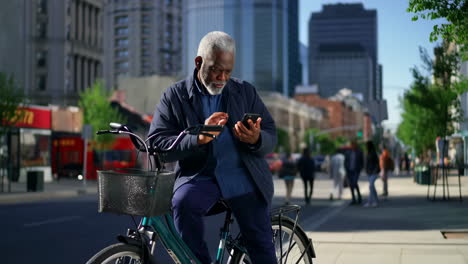 elderly african american man using smartphone on bicycle in city