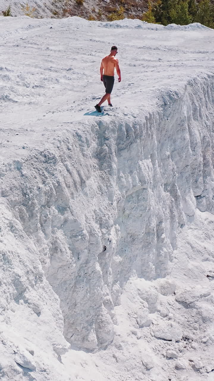 Bodybuilder in the mountains. Athletic man walking along the white rocky canyon. Sportsman in shorts doing exercises on white hill in summer. Aerial view. Vertical video