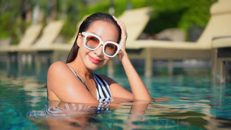 Señora-Encantada-Medio-Sumergida-En-La-Piscina,-Con-Gafas-De-Sol-Y-Traje-De-Baño-Sonriendo-Y-Mirando-A-La-Cámara