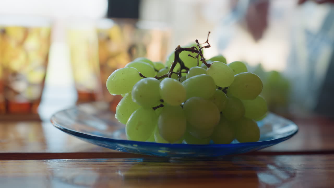 Close up of hand gently placing fresh green grapes on transparent blue glass plate, sunlight reflecting through honeycomb style drink glasses in background