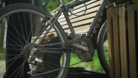 close-up of bicycle being placed between wooden bench and nearby structure, resting against bench while rider sits down, leans back, resting hands on legs, surrounded by lush green trees outdoor