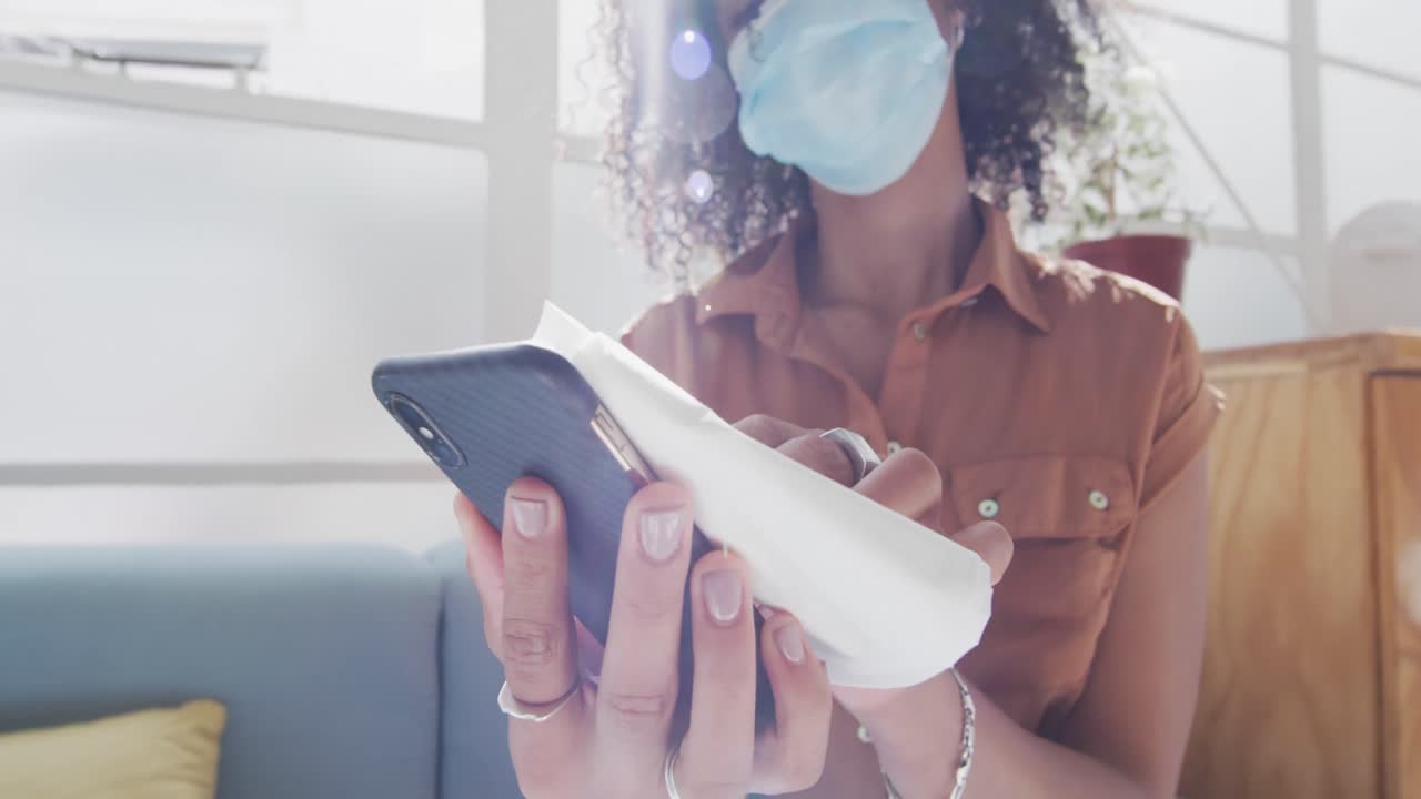 mujer con máscara facial limpiando su teléfono inteligente con un pañuelo
