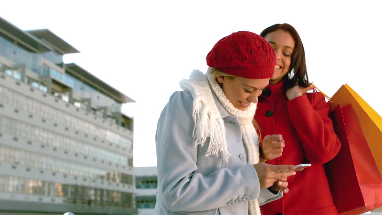 쇼핑백을 가진 예쁜 소녀들이 스마트폰을 보고 웃고 있습니다.