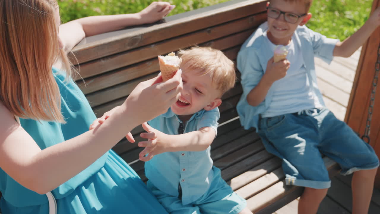 Caucasian family park bench laughter, sister playfully offers ice cream cone to little boy, older brother watches with smile, sunlit grass backdrop, summer weekend picnic vibe, candid sibling bond