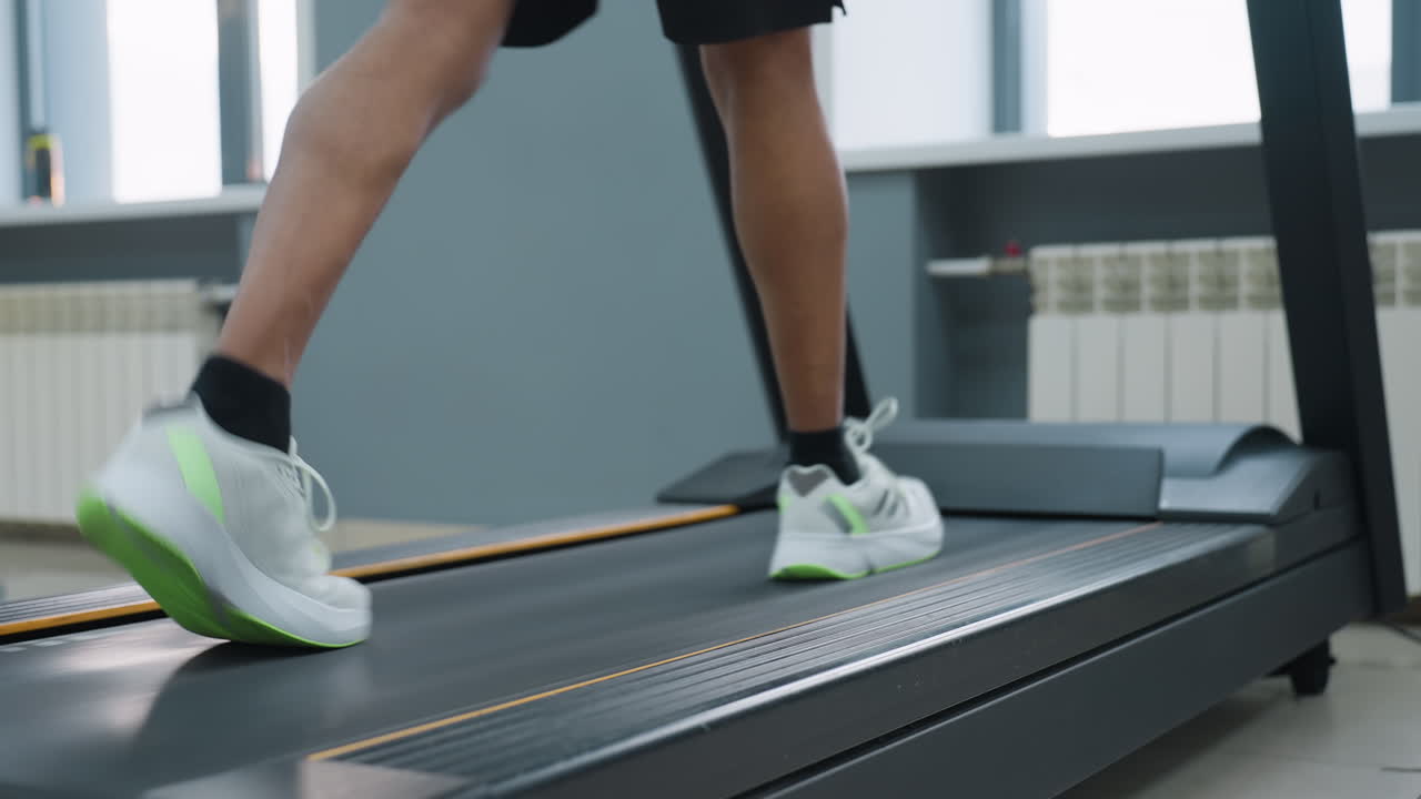 lower angle leg view of man in black shorts and neon sneakers briskly walking on treadmill belt in modern gym, light from window illuminating muscular calves, adjacent elliptical machine blurred