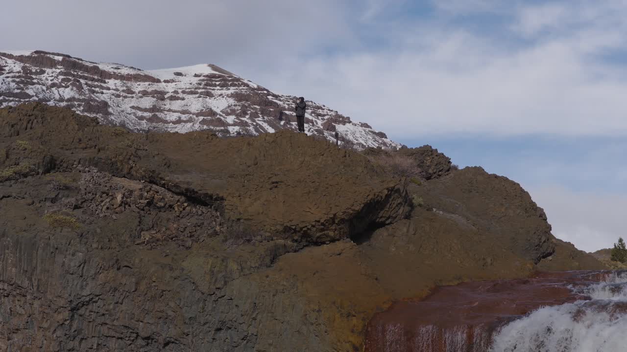 Man drinking yerba mate near Salto del Agrio waterfall with snow-covered mountains in the background