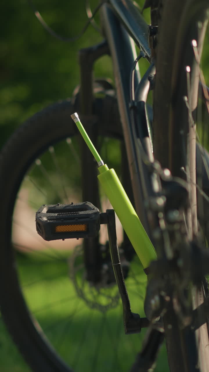 primer plano de la pierna y la mano de una mujer mientras quita una bomba de aire verde del área del pedal de su bicicleta, con un fondo verde exuberante de árboles y un edificio lejano borroso en el fondo
