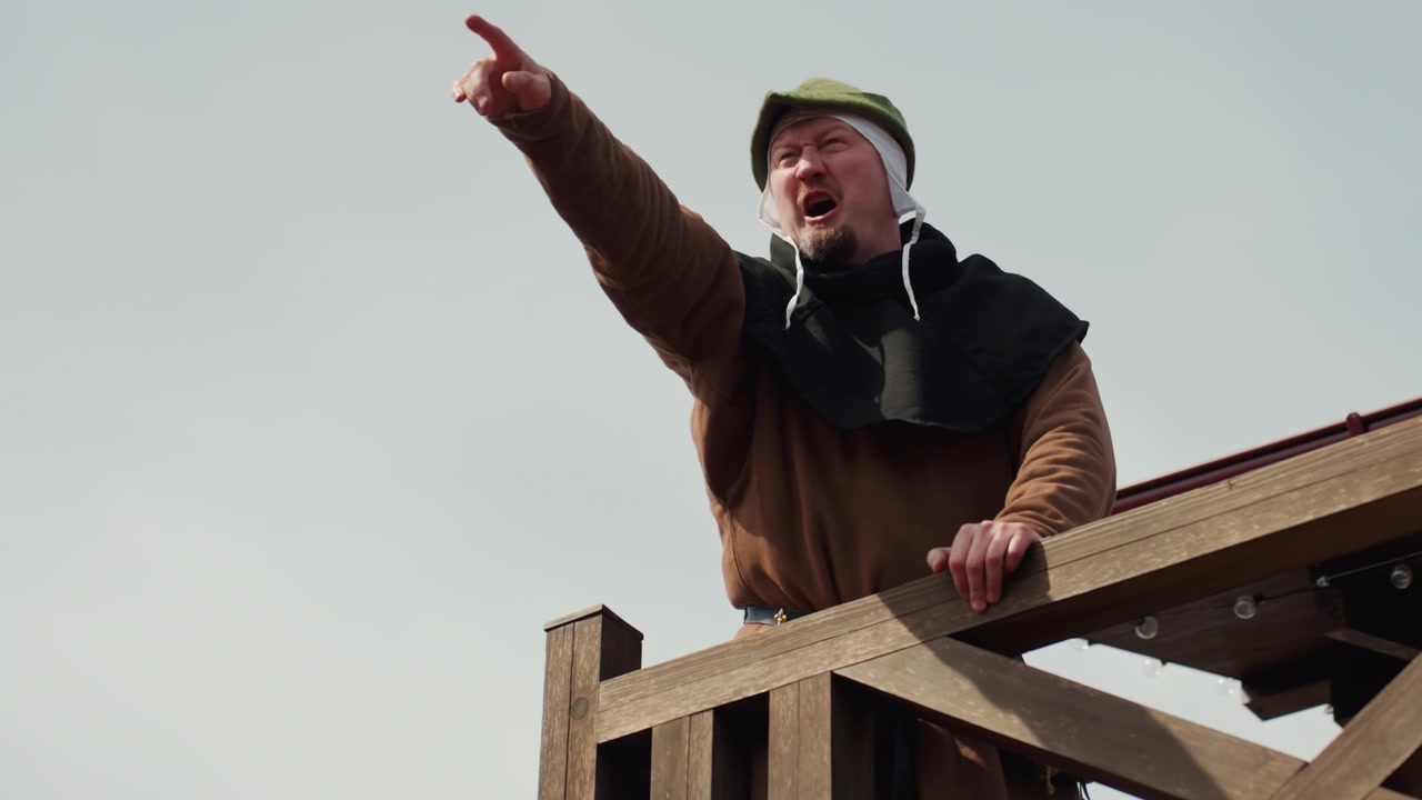 Un uomo anziano su una piattaforma di osservazione rustica in legno, assume pose pensierose mentre osserva la distanza, appoggiato alla ringhiera, riparandosi gli occhi, indicando, con una leggera brezza che gli muove il cappotto.
