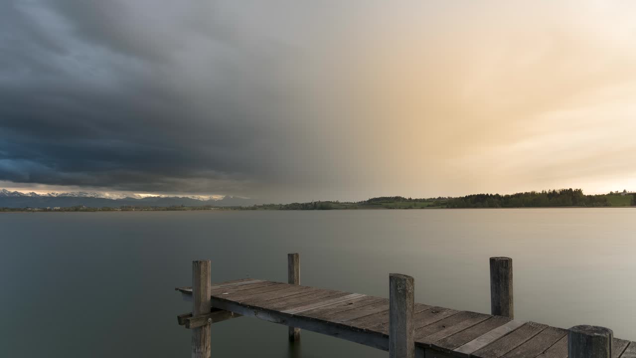 Storm clouds rise over a lake while rain, with the sun shining through. The mountains in the background of the natural landscape are illuminated and snow-covered. Shot on a jetty in spring. Time-lapse