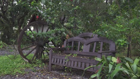 Pan-shot-of-a-secluded-park-bench-located-in-a-thick-dense-forest,-leafy-foliage-falling-from-the-trees-above-covering-the-seating-area-due-to-the-change-in-season