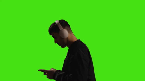 joven con auriculares inalámbricos y transmisión de música contra la pantalla verde con iluminación de baja clave caminando a través del marco