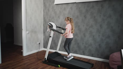 working out session of young girl on treadmill walking carefully hands on handles focused on balance and posture in home workout space with textured grey wall wooden floor and pink beanbag visible