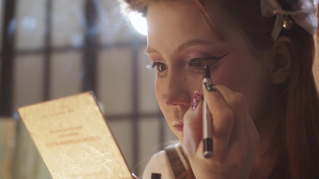 A cute girl doing makeup drawing dramatic winged eyeliner looking in a small mirror