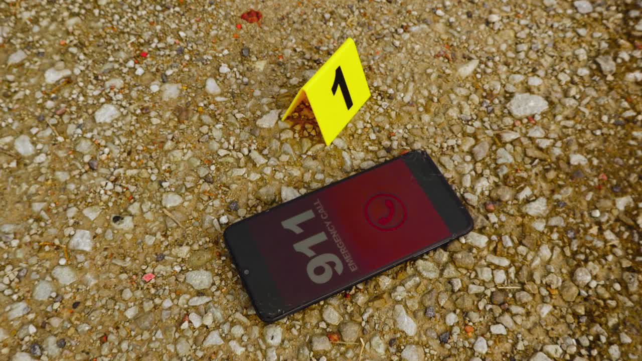 Top down view of a smartphone or phone that is dialing and calling 911 emergenvy number next to a yellow police evidence or crime scene marker on brown colored gravel outdoors on a road during day