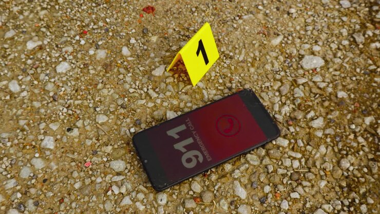 Top down view of a smartphone or phone that is dialing and calling 911 emergenvy number next to a yellow police evidence or crime scene marker on brown colored gravel outdoors on a road during day