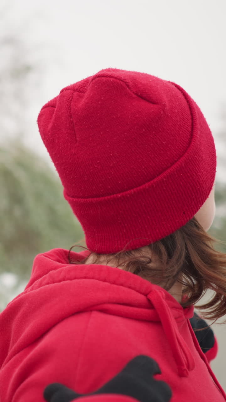 dama con capucha roja con las manos colocadas sobre los hombros gira el brazo y gira lentamente la cabeza hacia la izquierda en un parque cubierto de nieve, el fondo presenta árboles helados, bancos y una atmósfera de invierno serena