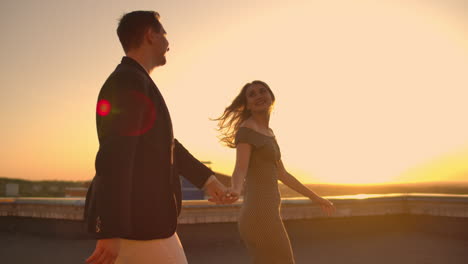 Young-couple-holding-hands-walking-woman-leading-boyfriend-the-roof-of-the-building-at-sunset-POV-travel-concept.-Carefree-free-lovers-run-on-the-roof-laughing-and-smiling