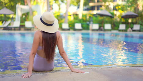 Una-Mujer,-Con-Traje-De-Baño-Y-Sombrero-Panamá,-De-Espaldas-A-La-Cámara,-Se-Sienta-En-El-Borde-De-La-Piscina-Y-Se-Relaja