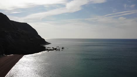 Antenne-Vom-Leeren-Seatown-Strand-Gegen-Ruhiges-Wasser-Mit-Der-Silhouette-Des-Klippensturzes-Im-Hintergrund