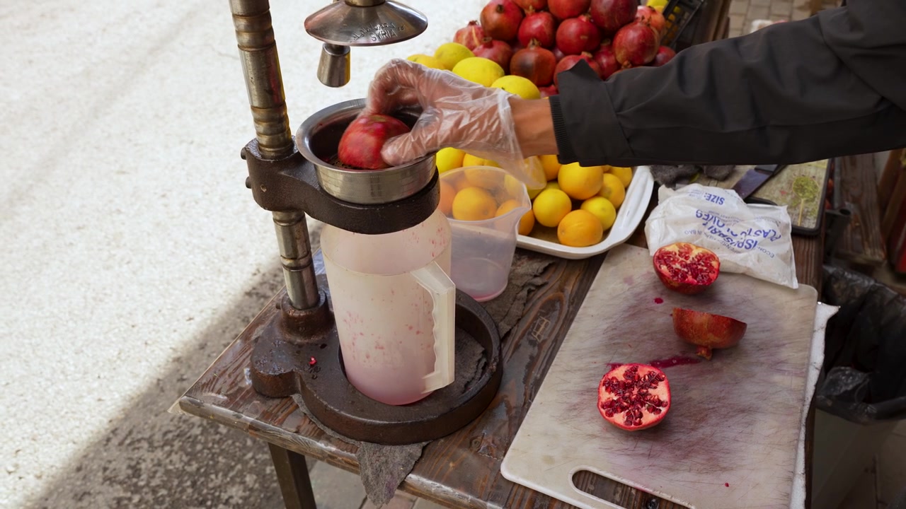 Uomo che spreme melograni freschi per farne succo in un mercato giordano locale usando una pressa manuale in metallo, inquadratura statica.