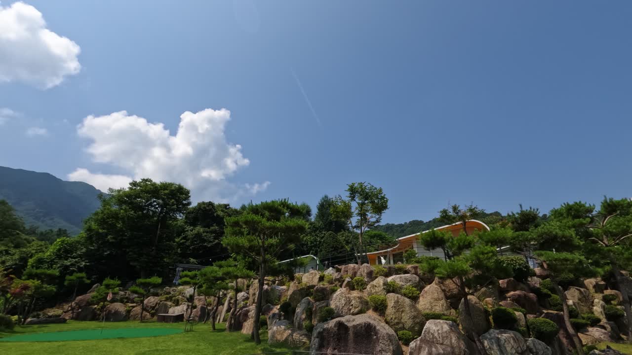 A beautiful time-lapse on a clear sunny day at a luxury glamping resort in Gapyeong, showing clouds moving across a blue sky above a landscaped rock garden and mountain