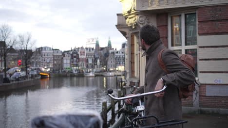 Hombre-Elegante-Admira-La-Vista-Del-Canal-De-Amsterdam-En-Un-Borde-De-Barandilla-Cerca-De-La-Bicicleta-Bloqueada