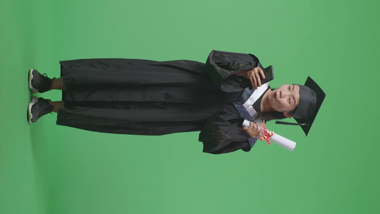 Laureata asiatica a figura intera in tocco e toga con diploma, che guarda lo smartphone ed esclama "wow" su sfondo green screen in studio