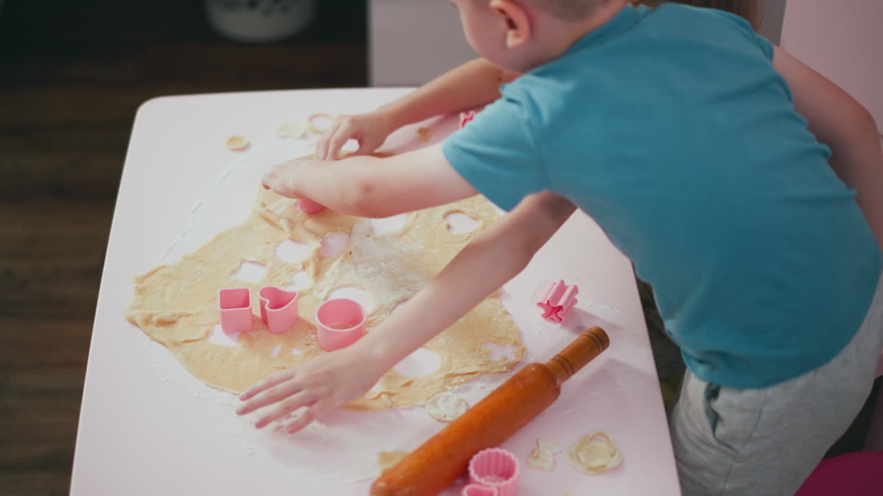 Aerial view of young boy trying to cut out shape on dough using cutter while girl reaches out for flour and boy prepares to sit back on chair during fun kitchen baking activity