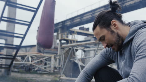 tired caucasian male boxer having rest near a punching bag during boxing training outdoors an abandoned factory on a cloudy morning