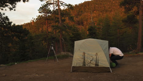Man-with-telescope-and-tent-in-forest-at-sunset,-long-shot