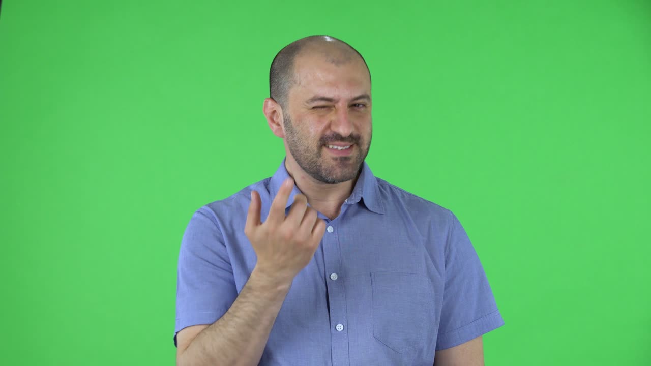 Portrait middle aged men looking at the camera playfully waves his hand and shows gesture to come here and winks. Balding male with beard in blue shirt posing on green screen in the studio. Close up