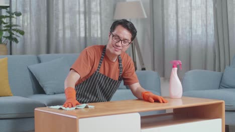 집에서 스프레이로 테이블을 청소하는 동안 카메라를 향해 미소 짓는 아시안 남성 집주인