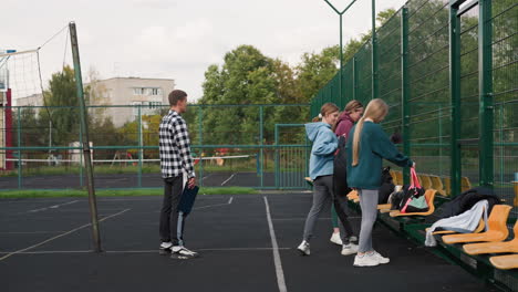 coach holding performance book patiently watches as students place bags on bench in volleyball court, outdoor sports setting with trees and building in background