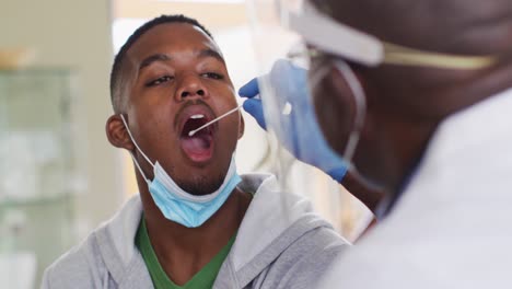 african american senior male doctor giving covid test to male patient in home, wearing face masks