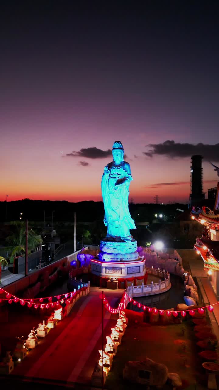 This vertical video showcases a Buddhist statue glowing in shifting colors under a mesmerizing sunset sky offering a serene and visually captivating scene of cultural and spiritual harmony