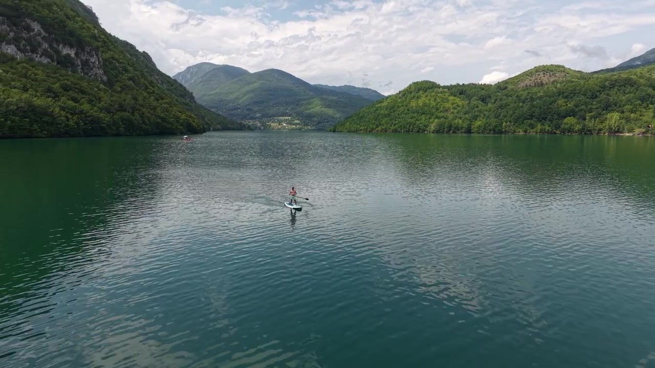 Ripresa aerea con drone di un uomo su paddle board in un lago verde con riflessi del cielo e delle colline verdi. Perucac, Serbia
