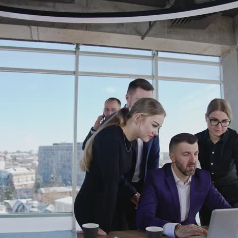 Group of colleagues stand around the boss with a laptop. Other businessman has a telephone conversation. Team working in a spacious office