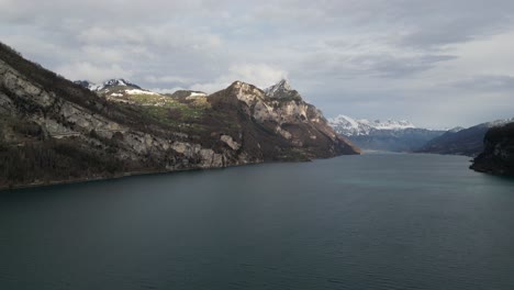 Dolly-above-lake-as-sunlight-breaks-between-clouds-illuminating-sheer-cliffs-and-leafless-trees