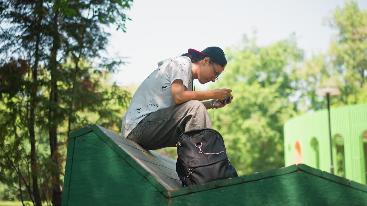 젊은 남자가 턱에 앉아 잠시 휴식을 취하며 휴대폰을 확인하고 있고, 앞쪽에는 나뭇잎이 장면을 감싸고 있으며, 배경에는 녹색 정자가 있고, 옆에는 배낭이 있으며, 따뜻한 햇살이 대비를 이루는 거리 사진 작가의 모습
