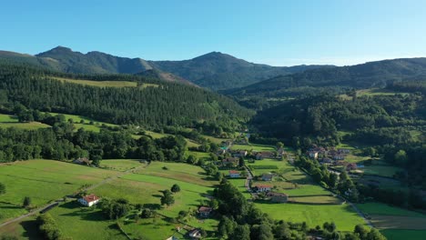 drone-flight-in-a-valley-seeing-a-village-with-its-white-houses-with-red-roofs,-crop-fields-and-their-roads-with-pine-forests-with-mountains-in-the-background-in-summer-at-sunset-iCantabria-Spain