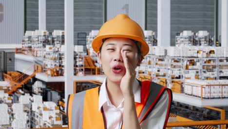 close up of asian female engineer with safety helmet yelling with hand over mouth while standing in the warehouse with shelves full of delivery goods