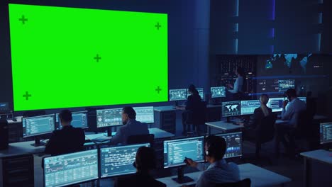 team of professional computer data science engineers work on desktops in control and monitoring room. big display has a green screen mock up, others showing charts, graphs and technical statistics.