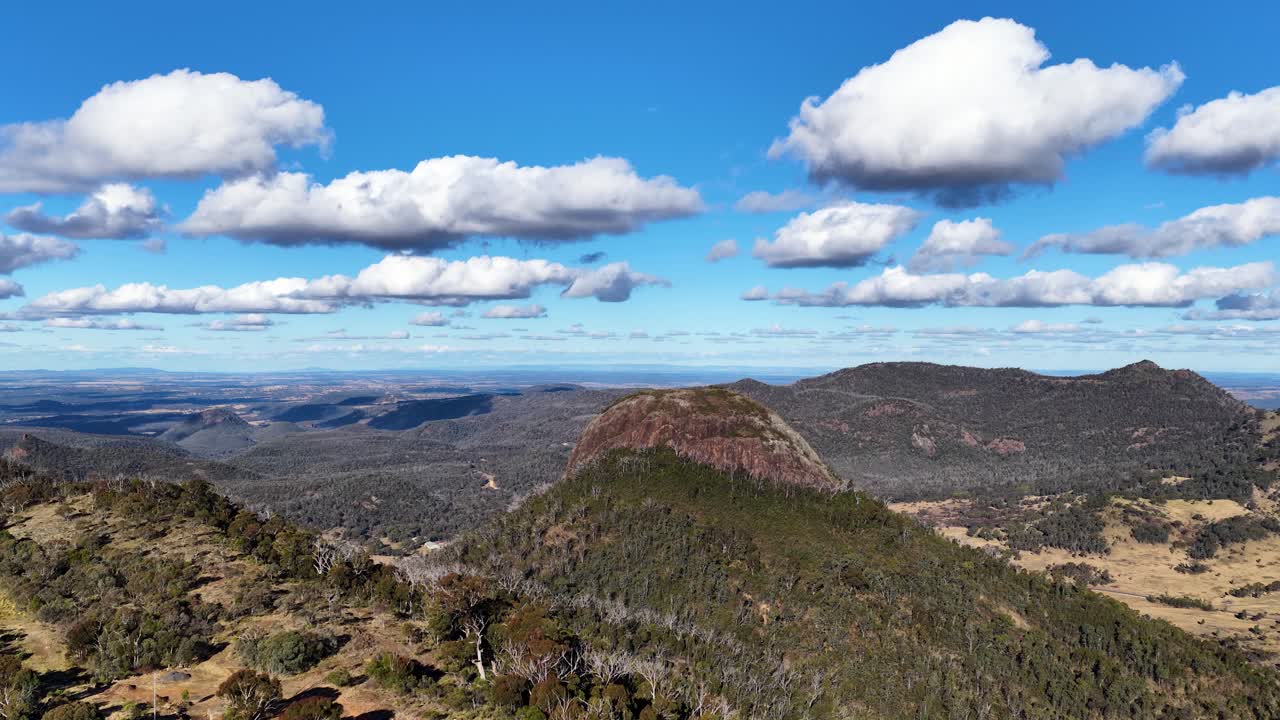 Drone ascends above forested hills and rocky peaks in Warrumbungle National Park, revealing expansive views under bright daylight and scattered clouds