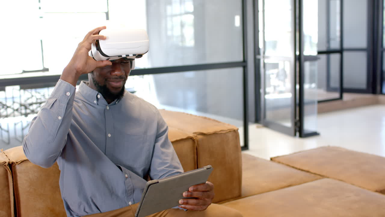 Using VR headset, man holding tablet while sitting on couch in office
