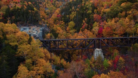 Un-Joven-Con-Una-Chaqueta-Amarilla-Explora-Un-Puente-De-Vías-De-Tren-En-La-Ladera-De-La-Montaña-Durante-El-Otoño-Con-Follaje-De-Otoño-En-El-Fondo