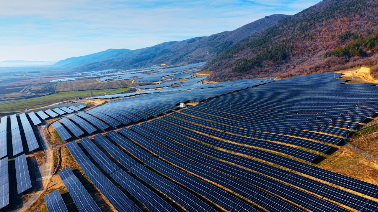 山岳地帯の大規模太陽光発電所を空撮。ブルガリアのベリラソーラーパークにて、澄み切った青空の下、丘陵地帯に設置された青いソーラーパネルの列。