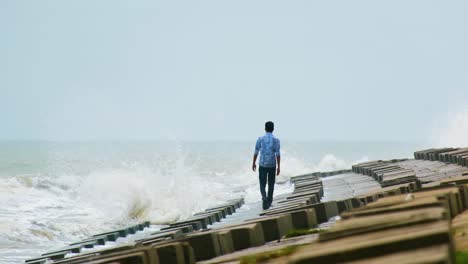 man appearing to be depressed and lonely walking at sea shore in slow motion against the crushing waves on a concrete breakwater slope