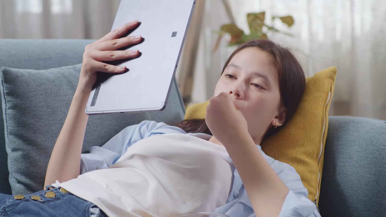 Close Up Of Asian Teen Girl Enjoys Using Tablet And Eating Snacks While Lying On Sofa In The Living Room At Home